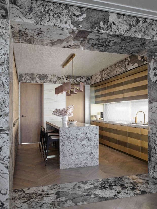 A refined contemporary kitchen viewed through a dramatic stone-clad opening. The space features a large marble or natural-stone island, matching stone surrounds and splashback, warm timber herringbone flooring, timber cabinetry with horizontal bronze-toned accents, a timber door, a textured light ceiling, and a sculptural pendant over the adjacent dining area. A window with a layered Roman blind and a brass tap bring additional warmth and detail.