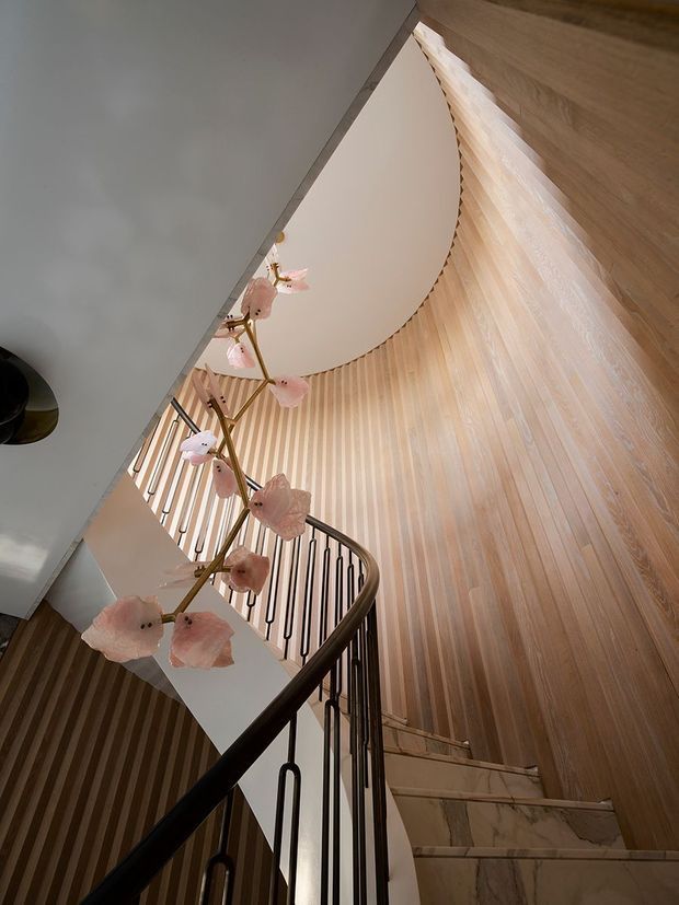 A dramatic upward view of a sculptural curved staircase with pale marble treads, dark metal balustrades, and warm vertical timber wall panelling. A large circular ceiling opening and delicate pink floral pendant add a refined decorative focus.