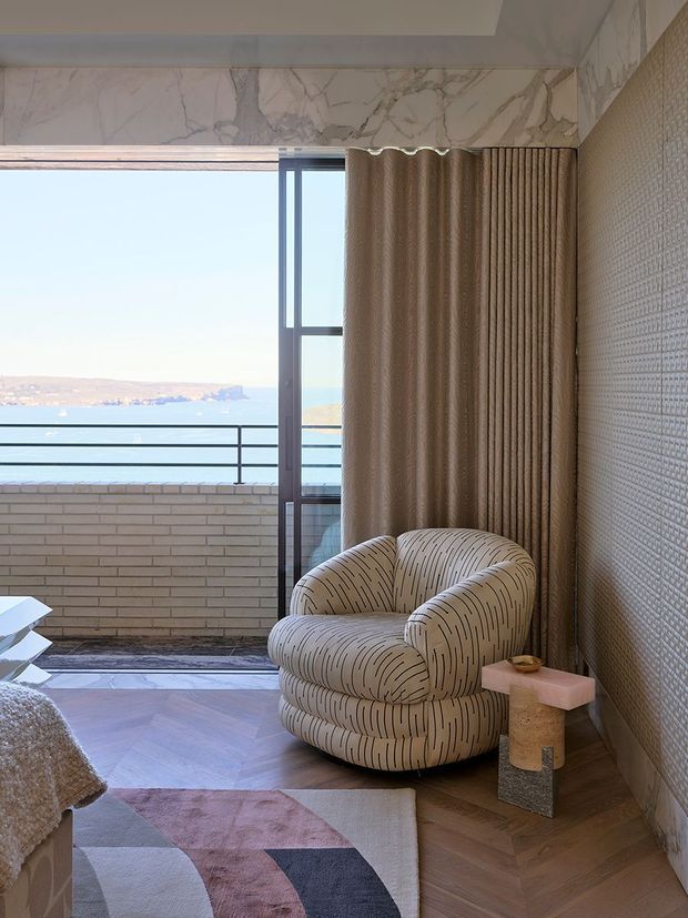 A refined living room corner with a sculptural upholstered lounge chair beside a small stone and pink-toned side table. Floor-to-ceiling glazing opens onto a balcony with expansive coastal water views. The room features warm herringbone timber flooring, textured wall covering, veined marble-look wall panels around the window, ribbed curtains, and a geometric area rug.