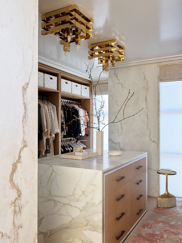 A luxurious walk-in wardrobe or dressing room with extensive open hanging storage, integrated shelving, and a central island dresser clad in dramatic white marble with grey veining. Warm wood drawer fronts, brass geometric ceiling chandeliers, decorative wall moulding, a patterned window shade, and a small sculptural side table create an opulent, refined interior.