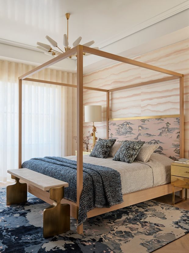 A refined bedroom featuring a timber four-poster canopy bed with blue and neutral bedding, patterned decorative pillows, a sculptural wooden bench, brass-toned bedside furniture, and a large abstract floral rug. The room has sheer curtains, ornate ceiling moulding, a sculptural pendant light, and a dramatic wave-patterned feature wall with a framed scenic panel. The overall look is layered, elegant, and contemporary with vintage-inspired details.