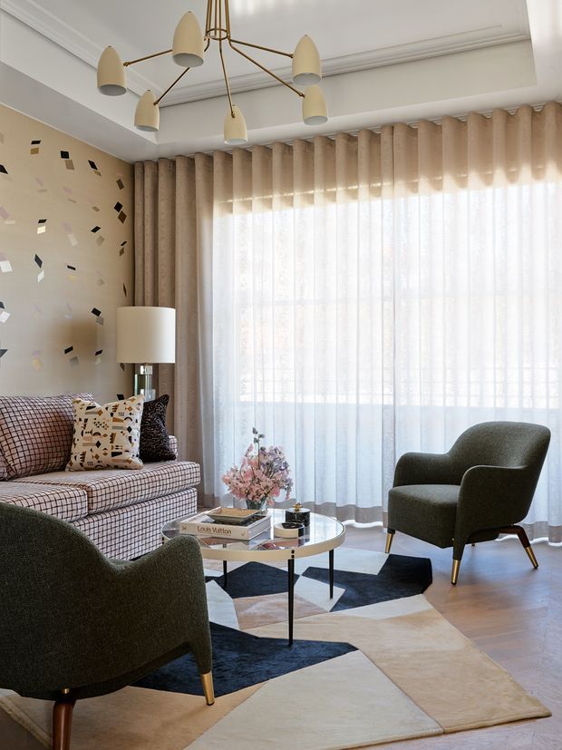 A refined living room with a large floor-to-ceiling window dressed in layered sheer and textured beige curtains. The room features a patterned upholstered sofa, two dark green lounge chairs, a round coffee table, a geometric area rug, a sculptural brass-and-cream pendant light, decorative cushions, and a floral arrangement. The palette is warm and muted with contemporary classic detailing, including ceiling cornices and wall panelling-like trim.