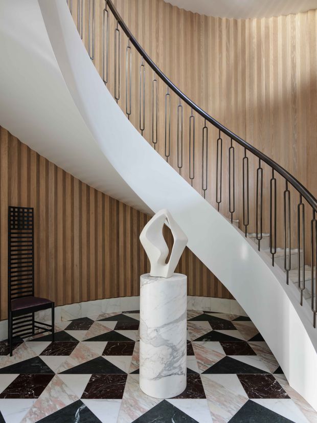 A refined stairway foyer with a sweeping white curved staircase and slim black metal balustrade. The walls are clad in vertical timber battens, while the floor features a geometric checkerboard pattern of black, white, and blush-toned stone or marble. A white sculptural artwork sits on a cylindrical marble pedestal, with a dark wood and black chair at the side.