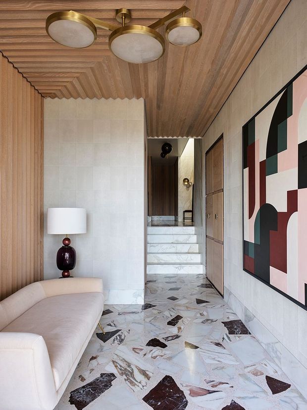 A refined residential hallway or entry passage with a sculptural cream sofa, a dark burgundy glass table lamp, geometric abstract artwork, warm timber wall and ceiling panelling, pale stone wall tiles, and a dramatic terrazzo floor. The corridor leads toward a short flight of marble-clad stairs, with brass and opal-glass ceiling lighting adding a luxe accent.