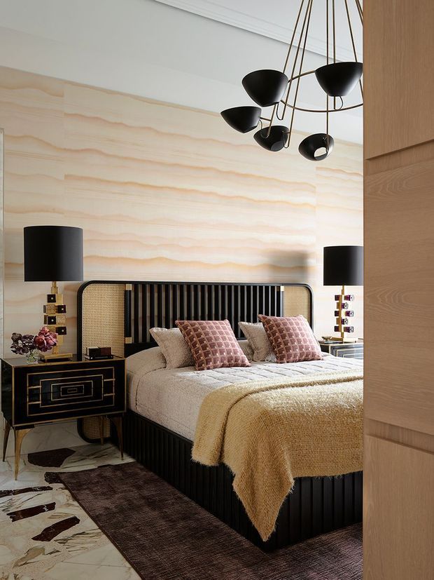 A sophisticated bedroom viewed through a partially open timber door. The room features a low black fluted bed with a rounded cane-panel headboard, layered neutral bedding, mauve patterned cushions, and a textured tan throw. A dramatic black multi-arm pendant hangs overhead, while matching black bedside lamps sit on geometric black-and-gold nightstands. The feature wall is clad in softly veined pale stone or timber-like panels, complemented by a marble-patterned floor, dark rug, and warm wood joinery. The styling is luxurious, refined, and strongly Art Deco influenced.