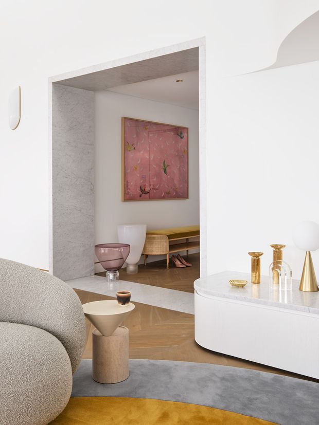 Refined contemporary living room with a curved cream boucle sofa, sculptural side table, grey and ochre area rug, and a rounded white built-in media or display cabinet. A large stone-clad portal opens to a secondary seating or entry area with timber flooring, a low console, artwork, and sculptural decor. The palette is soft white, blush pink, warm timber, grey, and brass, with curved architectural detailing and a polished, gallery-like atmosphere.