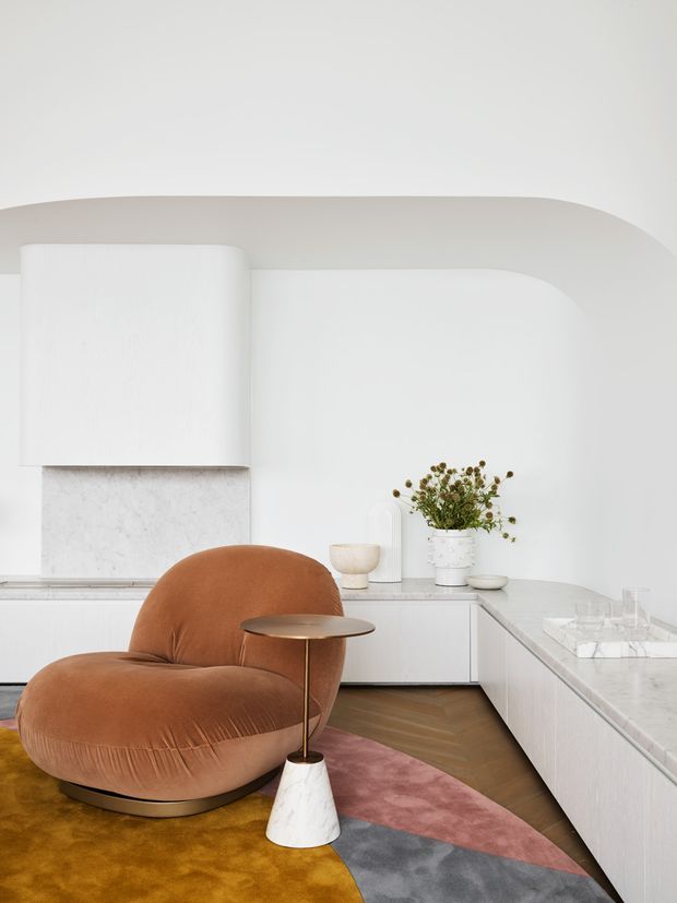 A refined contemporary kitchen and living corner with curved white architectural detailing, white cabinetry, and a pale stone island and benchtop. A sculptural rust-brown upholstered lounge chair sits beside a slender brass and marble side table, with a multicoloured abstract rug on a herringbone timber floor. The space is bright, minimalist, and softly rounded, with a small floral arrangement and decorative objects on the rear counter.