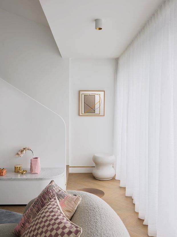 Bright, softly styled contemporary interior corner with white curved built-in joinery and a pale stone-like countertop, floor-to-ceiling sheer curtains, light timber flooring, a sculptural white side table, framed abstract artwork, and a rounded boucle lounge chair with patterned cushions. The space has clean curved forms, warm neutral finishes, and abundant diffused daylight.