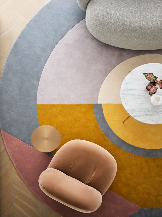 Overhead view of a stylised lounge seating arrangement on a large geometric area rug. A rounded taupe upholstered chair and a rust-toned curved lounge chair surround a small round metallic side table, while a circular marble-topped coffee table sits over concentric and segmented rug shapes in mustard, mauve, grey, burgundy and beige. Light timber parquet flooring is visible around the rug.
