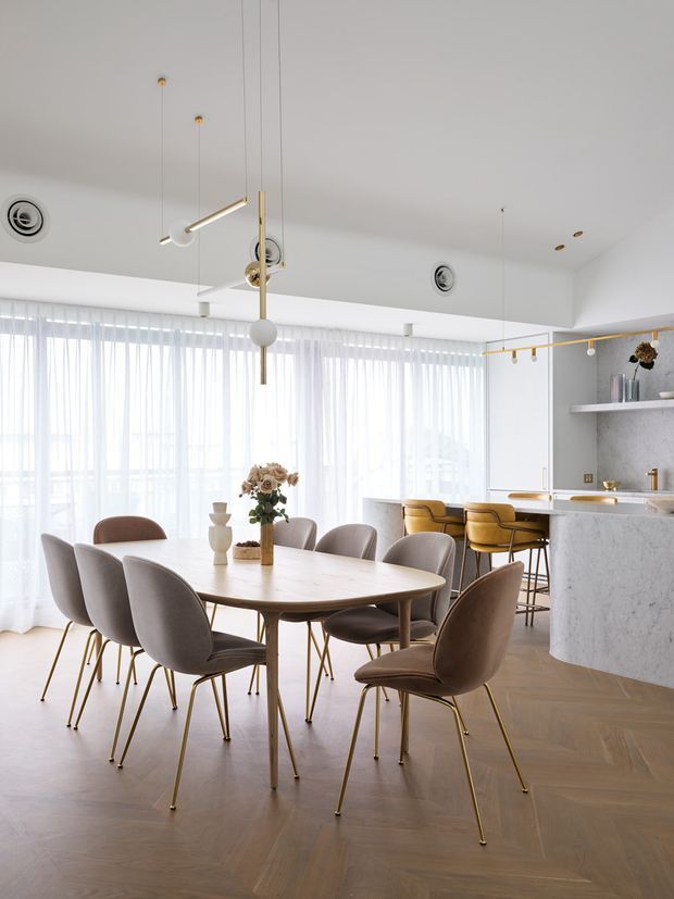 Bright open-plan dining and kitchen interior with a large oval dining table surrounded by upholstered chairs, floor-to-ceiling sheer curtains, sculptural pendant lighting, and a curved marble kitchen island with bar stools. The space has warm herringbone timber flooring, pale walls, and refined brass accents.