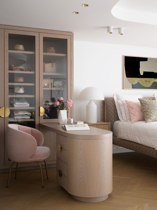 A refined bedroom interior featuring warm light-wood cabinetry with glazed display doors, a curved wood bedside or dressing console, a blush-pink upholstered chair, and a neatly dressed bed with layered neutral and pink textiles. The room has white walls and ceiling, timber flooring, brass-toned hardware and lighting, sculptural bedside lighting, and contemporary abstract artwork. The overall styling is soft, elegant, and modern with rounded forms.