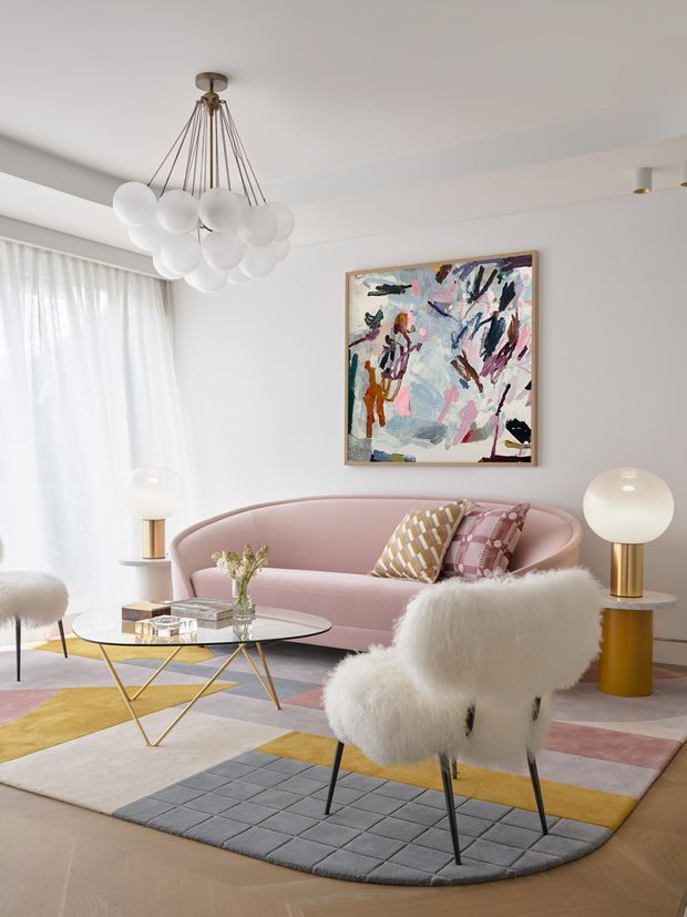 A bright, refined living room with a curved blush-pink sofa, sculptural white globe pendant, glass coffee table with gold-toned legs, fluffy white armchairs, and brass-accented floor lamps. An abstract artwork hangs on the white wall, while a multicoloured geometric rug adds mustard, pink, cream, and grey accents over a light timber floor.