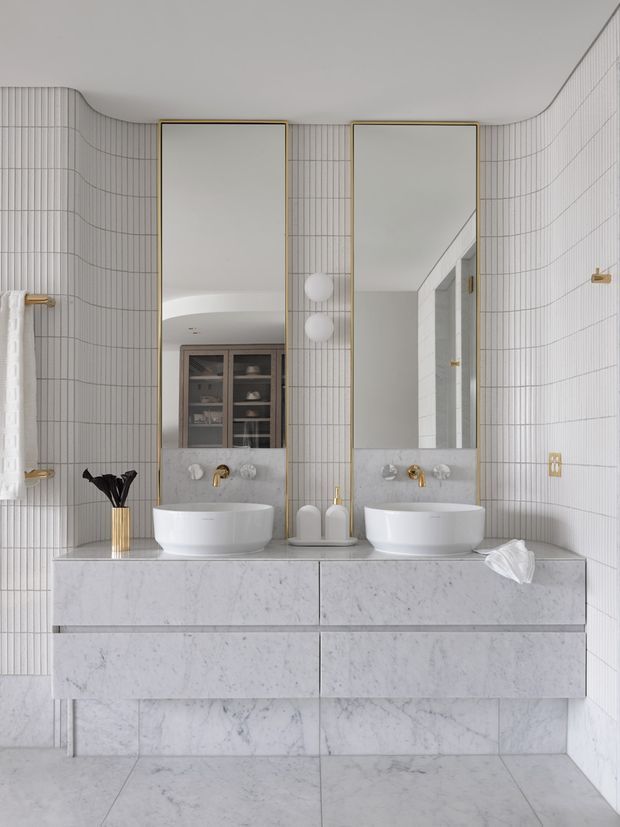 Bright, refined double-vanity bathroom with a symmetrical wall-mounted marble vanity, two white vessel basins, tall gold-framed mirrors, white vertically lined wall tiles, and brass fittings. A curved tiled wall adds a soft architectural detail, while a reflected built-in cabinet is visible in the left mirror.