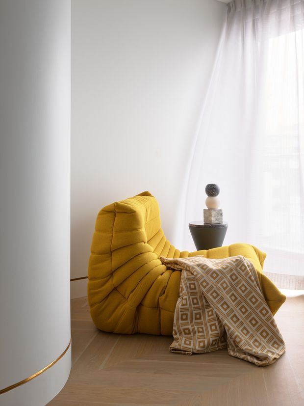 A bright, softly styled corner featuring a sculptural mustard-yellow upholstered lounge chair beside sheer white curtains. A patterned beige-and-white throw drapes over the chair and spills onto the light timber floor, with a small dark pedestal side table and decorative objects nearby. A curved white architectural wall with a slim brass accent detail frames the scene.