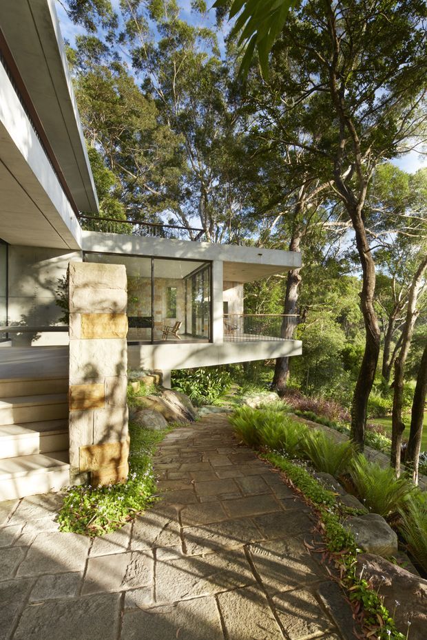 Exterior view of a modern, elevated concrete-and-glass house set among mature trees and landscaped gardens. A paved stone pathway leads beneath a projecting terrace and glazed living area, with broad concrete stairs, a stone-clad structural column, dark railings, and extensive surrounding bushland.