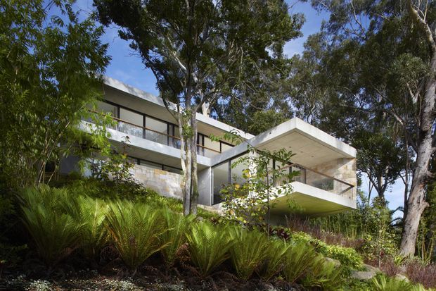 A modern, multi-level residence set within a heavily landscaped bush setting. The house features strong horizontal lines, flat projecting roof planes, extensive glazing, pale masonry walls, and sheltered balconies or terraces integrated into the sloping site.
