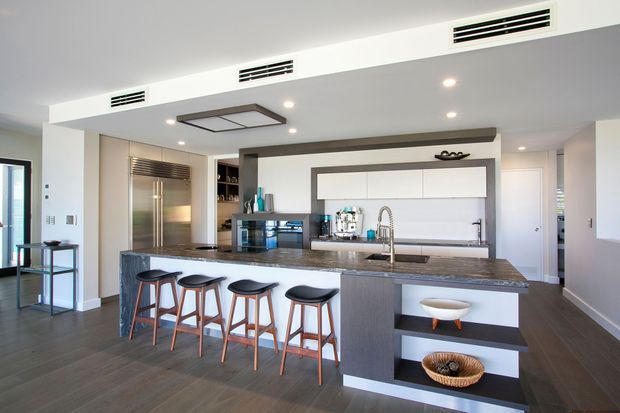 Contemporary open-plan kitchen with a large stone island, four timber-and-black stools, integrated stainless-steel refrigerator, white handleless cabinetry, dark timber-look flooring, recessed lighting, and a breakfast-bar display niche. The space is bright and streamlined with neutral finishes and strong indoor-outdoor glazing at the left.