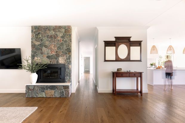 Bright open-plan residential interior with warm timber flooring, white walls and ceiling, and a prominent rough natural-stone fireplace surround with a built-in black firebox. A traditional dark timber wall mirror and console add character, while the space opens toward a contemporary kitchen or living area in the background.