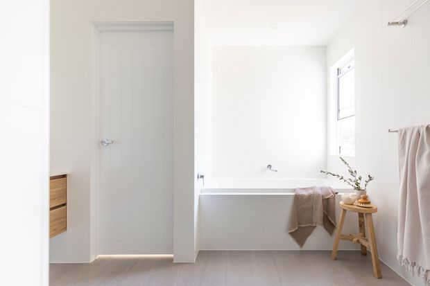 Bright, minimalist bathroom with a white built-in bathtub beneath a window, pale timber-look flooring, white walls, a light grey interior door, and a small wooden stool with towels and a plant. A partial timber vanity or drawer unit is visible at the left edge.