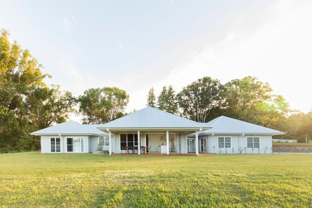 Wide exterior view of a low, single-storey residence set behind an expansive lawn. The home has a symmetrical, light-coloured facade with multiple gabled roof sections, a central covered outdoor entertaining area, white-framed windows and doors, and a leafy landscaped setting.