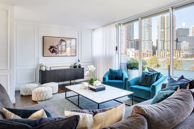 Bright, elegant living room with white decorative wall panelling, full-height corner glazing, a dark timber sideboard, large sectional sofa, teal armchairs, ottomans, and a rectangular coffee table. A dense modern city skyline and water are visible through the windows.