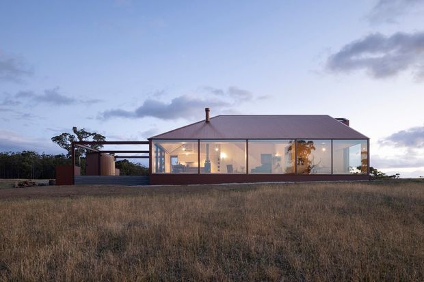A low, single-storey contemporary house set in an open grassy landscape at dusk. The building has a simple rectangular form, a broad hipped roof, extensive floor-to-ceiling glazed walls, and a covered outdoor structure or carport extending from the left side. Warm interior lighting is visible through the glazing, while the exterior is finished in muted dark and neutral tones.