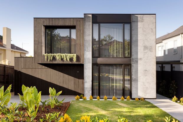 Front exterior view of a contemporary two-storey residence with a strongly rectilinear facade. The building combines dark vertically ribbed cladding, large black-framed glazed openings with vertical blinds, and pale textured concrete or stone-like wall panels. A small upper-level planter balcony, landscaped front garden, lawn, and concrete path are visible, with neighbouring homes on either side.