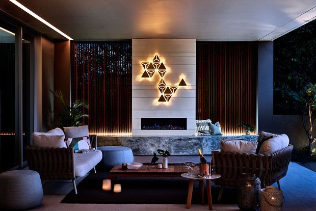 A sophisticated contemporary living room with a linear fireplace set into a pale horizontal-panel feature wall, illuminated geometric wall art, dark vertical timber screens, and a large stone hearth. The room is furnished with deep brown upholstered lounge chairs, a pale blue sofa, and a low timber coffee table, with warm accent lighting and views to greenery through dark-framed glazing.