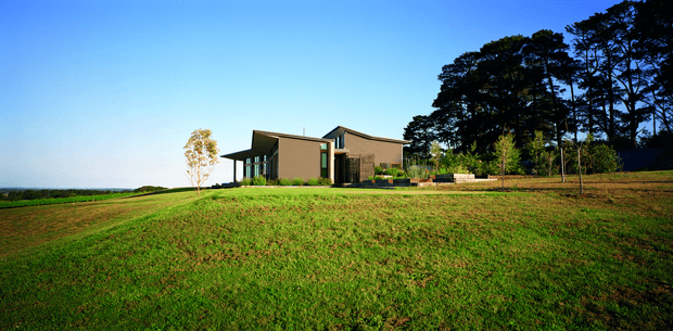 A low, contemporary single-storey house set within a broad landscaped lawn. The building has angular intersecting roof forms, dark window framing, and muted earthy exterior walls, with a dense stand of tall trees behind it and an expansive open horizon to the left.