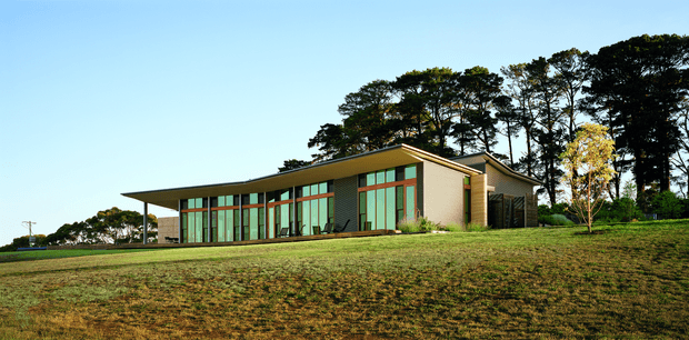 A low, single-storey contemporary building set within a broad landscaped lawn. The structure has a long glazed facade with timber-toned vertical framing, pale rendered walls, a shallow pitched roof, and a projecting flat-roofed entrance or canopy. Mature trees form a backdrop around the site.