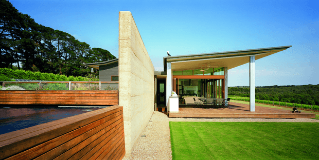 A contemporary single-storey pavilion-style house set in a landscaped rural environment. The image shows a broad timber-clad raised deck and pool or water feature in the foreground, a tall raw concrete privacy wall, extensive floor-to-ceiling glazing, and a dramatic thin projecting roof canopy over an open outdoor living area. A bright lawn and surrounding green landscape are visible beyond.