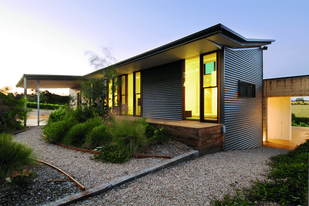 Single-storey contemporary house with a low-slung asymmetrical roof and corrugated metal cladding. Warm yellow light glows through tall vertical windows and glazed doors, contrasting with dark horizontal metal siding and a low brick base. A gravel pathway and landscaped native-style planting lead toward the entrance, with a covered outdoor structure extending to the left.