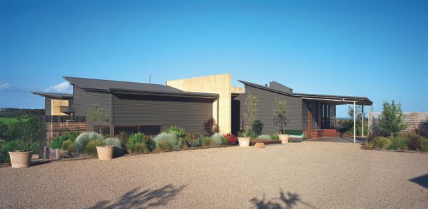 A contemporary single-storey residential building viewed from the front across a broad gravel forecourt. The low-profile composition has dark horizontal cladding, pale timber-toned wall sections, projecting mono-pitched roof planes, extensive glazing, a covered entry or veranda, and landscaped planting beds. The setting appears open and semi-rural, with distant landscape visible beyond the property.