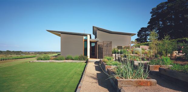 A compact contemporary single-storey residence set within a landscaped rural garden. The home has horizontally clad grey-brown walls, projecting asymmetric roof planes, large glazed openings, and a timber entry door. A gravel or concrete path leads through raised timber garden beds toward the entrance, with an expansive lawn and open agricultural landscape beyond.