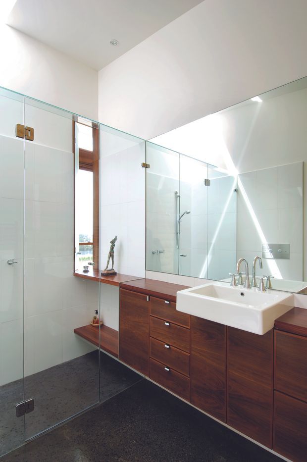 Bright contemporary bathroom with a large frameless glass walk-in shower, dark polished concrete flooring, white tiled walls, extensive mirrored surfaces, and a long dark timber vanity with integrated drawers and a white basin. Natural light enters through a narrow timber-framed window, creating strong reflections across the mirrors.