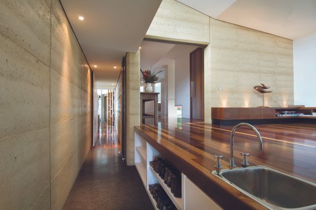 A refined open-plan kitchen and circulation space with a long polished timber island and inset stainless-steel sink in the foreground. The kitchen opens toward a living area with low timber joinery and sculptural decor. A long hallway runs alongside a textured exposed concrete wall, while recessed ceiling lighting and warm timber flooring create a sophisticated, linear interior.