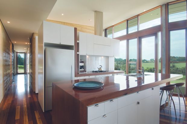 A bright, open-plan contemporary kitchen with a large timber island, white integrated cabinetry, stainless-steel appliances, and a reflective pale splashback. Full-height and clerestory-style glazing with dark timber frames opens the kitchen to expansive rural views. Dark timber flooring extends into the hallway, while slim bar stools provide seating at the island. The space has clean lines, abundant natural light, and a warm material palette.