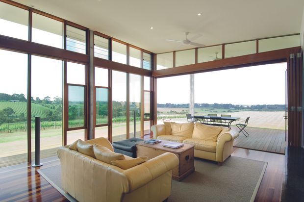 A bright, spacious living room with high ceilings and extensive floor-to-ceiling timber-framed glazing overlooking an open rural landscape. Two cream leather sofas face a dark rectangular coffee table and timber trunk-style side table, with a large neutral rug over polished timber flooring. The room opens onto a timber deck furnished with an outdoor dining table and chairs. Ceiling fans, recessed lighting, and dark timber framing create a warm contemporary pavilion-like interior.