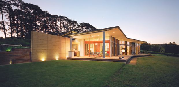 A dusk exterior photograph of a low-slung contemporary house set within a landscaped lawn and wooded site. The home has a projecting flat roof, extensive floor-to-ceiling glazing, warm interior lighting, timber and concrete-toned wall surfaces, and a broad covered terrace. The composition emphasizes the single-storey building form and its relationship to the surrounding garden and trees.