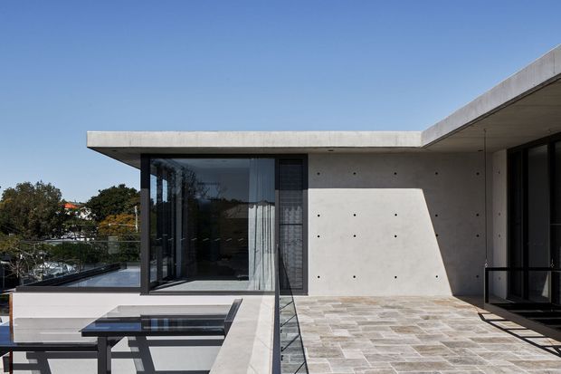Exterior view of a minimalist contemporary flat-roof residence, featuring exposed smooth concrete walls and roof slabs, expansive dark-framed glazing, a paved terrace, and a reflective pool or water feature in the foreground. The clean horizontal geometry and strong sunlight create a refined modern architectural composition.