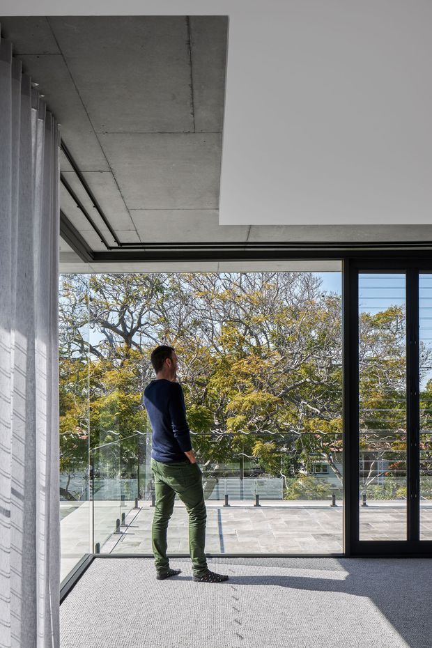 Contemporary interior opening onto a spacious terrace through full-height glazing. The room features a smooth exposed concrete ceiling, dark metal-framed sliding doors, pale wall surfaces, and grey carpet. A glass balustrade and leafy urban landscape are visible outside, with a person standing near the glazing.