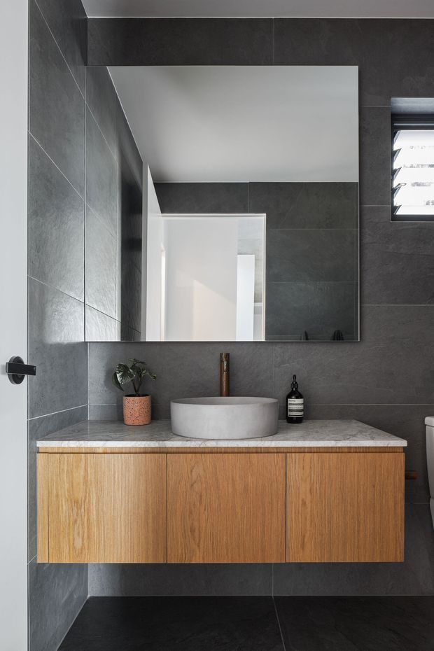 Contemporary bathroom vanity with a floating oak-toned timber cabinet, pale stone benchtop, round vessel basin, brushed metal tap, and a large frameless mirror. Dark charcoal large-format wall and floor tiles create a dramatic contrast with the warm timber joinery. A small high window provides natural light.