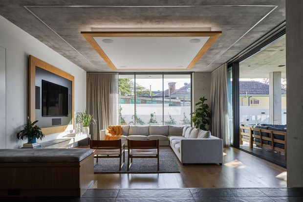 A refined contemporary living room opening onto a covered outdoor entertaining area through large dark-framed sliding glass doors. The room features a grey sectional sofa, timber-framed lounge chairs, a low dark timber media cabinet, sheer curtains, indoor plants, and a wall-mounted television framed by timber trim. The ceiling combines a textured concrete-look finish with a recessed white feature panel edged in timber, while warm timber flooring and abundant glazing create a bright, connected interior.