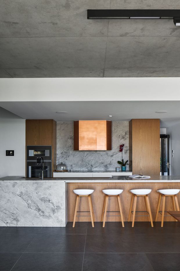 Contemporary kitchen with a large marble-look island and integrated breakfast bar, four timber stools, warm timber cabinetry, dark tiled flooring, and a dramatic copper-toned rangehood canopy. The rear wall features a grey veined stone or marble-look splashback, with built-in dark appliances and a concrete-look ceiling with linear lighting.