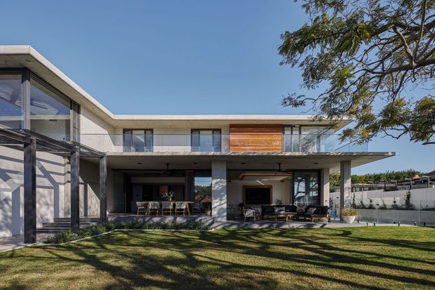 Exterior view of a contemporary two-storey residence with a flat roof, broad cantilevered upper floor, extensive glazing, concrete structural columns, and a covered outdoor living area opening onto a landscaped lawn. Timber wall cladding adds warmth to the otherwise pale concrete and glass facade.