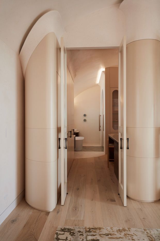 Warm, sculptural interior view through open white doors into a compact bathroom and adjoining vanity area. The space features curved organic walls and cabinetry, pale timber flooring, light neutral finishes, built-in storage, a toilet, and a softly lit arched ceiling and passage.
