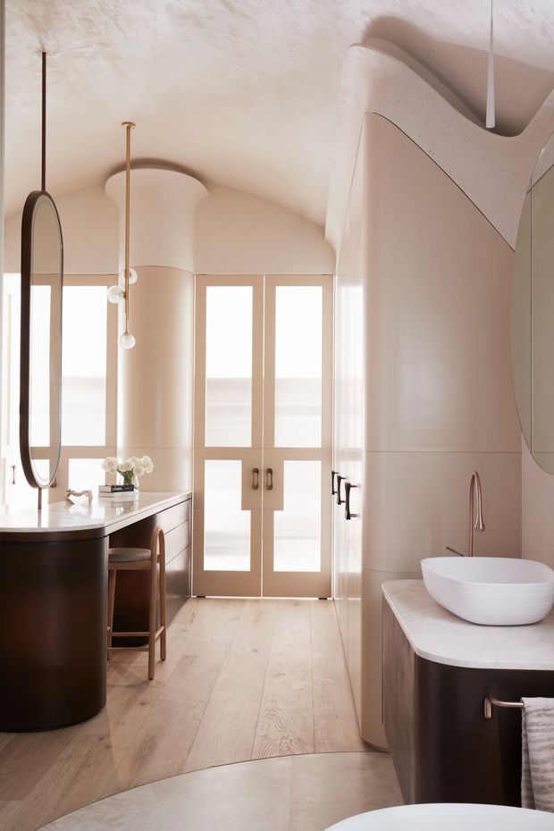 A refined, softly curved bathroom interior with a freestanding bathtub partially visible in the foreground, a sculptural double-curved vanity and basin on the right, and a rounded dark-wood vanity with an oval mirror on the left. Pale timber flooring, warm blush-beige plastered walls and cabinetry, fluted glass double doors, brushed brass hardware, and delicate pendant lighting create a calm, luxurious atmosphere.