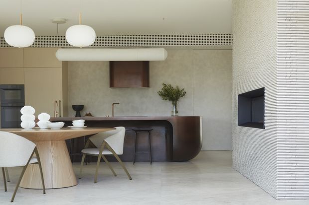 Contemporary open-plan kitchen and dining area with a sculptural dark island, pale timber cabinetry, curved dining table, upholstered chairs, globe pendant lights, and a textured linear feature wall containing a recessed fireplace or display niche. The space has a restrained neutral palette and refined minimalist detailing.