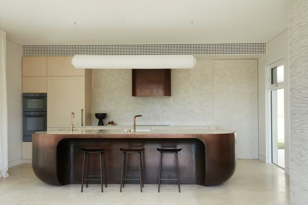 A refined contemporary kitchen with a large sculptural island featuring a dark rounded base and a light stone benchtop. Three stools sit at the island, with integrated cabinetry, built-in ovens, minimalist taps, a textured pale feature wall, and full-height glazing with blinds. The room has a clean, neutral, softly illuminated aesthetic.