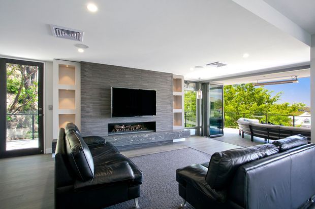 Bright contemporary living room with black leather sofas and armchairs arranged around a large grey rug, a wall-mounted television and linear fireplace set into a textured grey feature wall with integrated display niches. Full-height glazing and open glass doors connect the room to an outdoor balcony or terrace with leafy views.